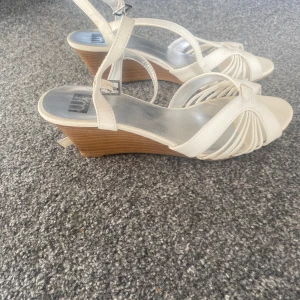 Vita sandaler med kilklack, strl 39 - Snygga vita sandaler med kilklack och öppen tå. Remmar i syntet skapar ett stilrent mönster över foten och runt vristen. Perfekta för sommarens alla tillfällen när du vill ha en fräsch och luftig look.