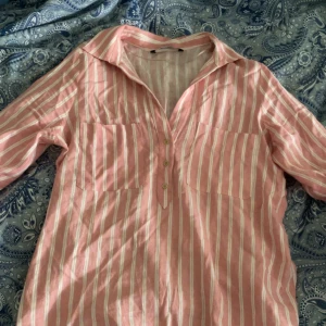 Rosa och vit randig skjorta från Zara - Säljer en rosa och vit randig skjorta från Zara i storlek M. Skjortan har trekvartsärmar med knapp, två bröstfickor och knappar framtill i guld. Tillverkad i ett lätt och luftigt linnematerial, perfekt för varmare dagar. Säljer pågrund av att den inte kommer till användning, har blivit använt ca 2 gånger.🤗