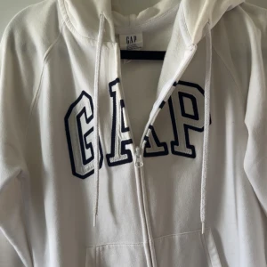Vit hoodie med dragkedja från GAP ! - Äkta vit hoodie från GAP i storlek XS med stor broderad logga framtill i mörkblått! Tröjan har en skön luva med snören, fickor samt dragkedja. Materialet är mjukt och består av bomull & polyester, funkar perfekt för alla tillfällen :)