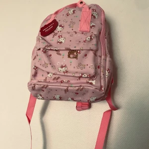 Hello Kitty rosa ryggsäck - Supergullig rosa ryggsäck med Hello Kitty-motiv och små färgglada detaljer. Väskan har justerbara axelband, ett stort huvudfack och en mindre ficka framtill med dragkedja. Perfekt för dig som älskar söta accessoarer och vill sticka ut med en ikonisk look.