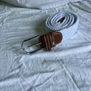 Vitt flätat bälte med bruna skinn-detaljer - Snyggt vitt flätat bälte med bruna skinn-detaljer och silvrigt spänne. Perfekt accessoar för att lyfta din outfit med en fräsch och stilren look. Bältet har en klassisk design med kontrasterande sömmar på skinnet.