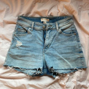 Ljusblå jeansshorts från H&M - Ljusblå jeansshorts från H&M med hög midja och råa, fransiga kanter. Shortsen har slitna detaljer både fram och bak, klassiska fem fickor och dragkedja med knapp. Bra skick och stretchigt material🤗