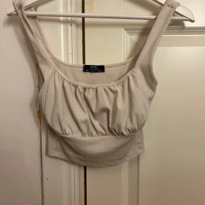 Ribbad linnetopp från Bershka - Superfin beige/ crämvit linnetopp från Bershka med breda axelband och ribbad struktur. Toppen har en lätt rynkad detalj framtill och är croppad i modellen. Perfekt att styla med jeans eller kjol för en chill look.