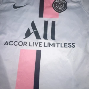 PSG Nike fotbollströja vit/rosa - Snygg PSG fotbollströja från Nike i vitt med rosa och mörkblå vertikal rand på framsidan. Tröjan har korta ärmar, rund hals och är tillverkad i lätt Dri-Fit material. PSG-logga och Nike Swoosh på bröstet samt sponsortryck på magen.