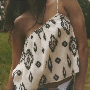 Zara  topp - Säljer en luftig vit oneshoulder topp med svart grafiskt mönster och smal axelrem med knyt. Toppen är croppad och har en avslappnad passform, perfekt för varma dagar. Materialet känns lätt och svalt mot huden.