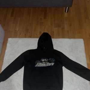 Svart hoodie Team Shooters - Svart hoodie med tryck av Team Shooters och pantermotiv på bröstet. Klassisk modell med huva, snörning och känguruficka framtill. Långärmad och i mjukt material, perfekt för chill dagar eller när du vill ha en sportig vibe.