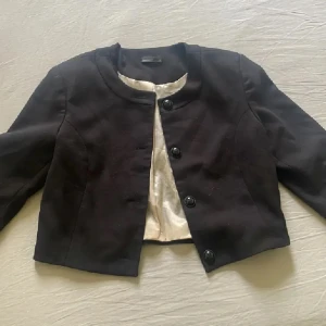 Svart croppad  kavaj med knappar - Stilren svart kavaj med rundad krage och tre stora knappar framtill. Jackan är croppad och har lång ärm samt ljus insida. Perfekt för dig som vill ha en klassisk och clean look.