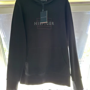 Svart hoodie Tommy Hilfiger S - Svart hoodie från Tommy Hilfiger i storlek S. Snyggt broderad logga på bröstet med röd ram och klassisk Hilfiger-flagga på ärmen. Tröjan har huva utan snöre och stor magficka framtill. Mjuk bomullskänsla och långärmad modell. Ungefär 500kr nypris via booztlet, köptes 2 år sedan men som inte kom till användning mycket. Var helt enkelt inte min stil