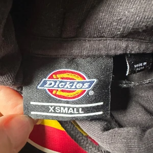 Svart Dickies hoodie XS - Svart hoodie från Dickies i storlek XS med stort färgglatt Dickies-tryck på bröstet. Tröjan har huva med snörning och en klassisk magficka. Perfekt för dig som gillar streetwear och vill ha en chill look.