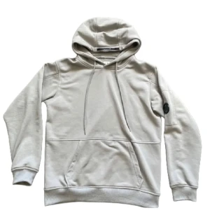 Beige hoodie från C.P. Company - Snygg beige hoodie från C.P. Company med klassisk huva och snörning. Tröjan har en stor magficka och den ikoniska linsen på ärmen. Tillverkad i mjuk bomull och passar perfekt för en chill och stilren look.