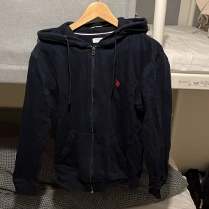Marinblå hoodie från Ralph Lauren - Marinblå hoodie från Ralph Lauren. med dragkedja, huva och snörning. Tröjan har en röd broderad logga på bröstet och två fickor framtill. Perfekt för en chill och stilren look.
