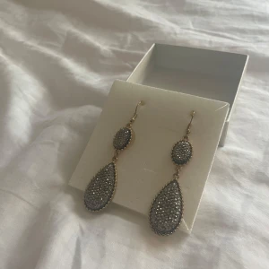 Glittriga droppformade örhängen - Säljer ett par snygga örhängen med droppformad design och glittriga silverfärgade stenar. Örhängena har guldfärgade kanter och krokar. Ursprungligen från Italien. Säljer då dem inte är min stil.