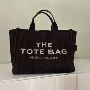 Svart Tore bah från Marc Jacobs - Cool svart väska från Marc Jacobs med tydlig vit text 'THE TOTE BAG' på framsidan. Väskan har två rejäla handtag och är tillverkad i slitstark bomull. Perfekt storlek för att få plats med allt du behöver och har en stilren, boxig form. Den är perfa till skolstarten