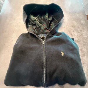 Svart hoodie från Polo Ralph Lauren - Asfrän Svart hoodie från Polo Ralph Lauren med dragkedja och klassisk gul broderad logga på bröstet. Tröjan är i Xs så passar dig som är runt 165-173. Fodret i huvan har rutigt mönster i grönt, rött och gult. Tröjan har känguruficka och är tillverkad i mjuk bomull. Perfekt för chill dagar eller streetstyle.