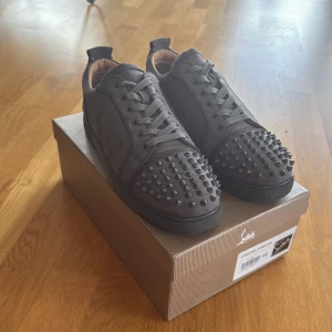 Christian Louboutin svarta sneakers med nitar - Grymma svarta sneakers från Christian Louboutin med mattsvarta nitar på tån och klassisk röd sula. Skorna har rund tå och är tillverkade i mocka.Perfekta för dig som vill sticka ut med en lyxig stil.