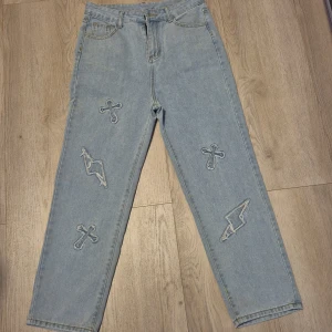 Ljusblå jeans med broderade kors, S - Unika ljusblå jeans med broderade kors och blixtar på både fram- och baksida. Raka ben och klassisk femficksmodell. Materialet är 65% bomull och 35% polyester för en skön känsla och bra passform. Perfekta för dig som vill sticka ut med coola detaljer.