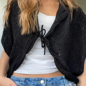 Svart stickad kofta med knyt - Svart stickad kofta med snygg passform och knyt framtill. Koftan har en luftig och grovstickad struktur som ger en avslappnad vibe. Perfekt att slänga över ett linne eller t-shirt för en chill look.