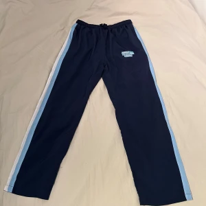 Norra Läktaren Malmö  - Mörkblå byxor med ljusblå och vita ränder längs sidorna. Byxorna har resår i midjan med snörning och ett broderat Norra Läktaren Malmö-märke på låret. Perfekta för dig som vill ha en sportig och bekväm look. 