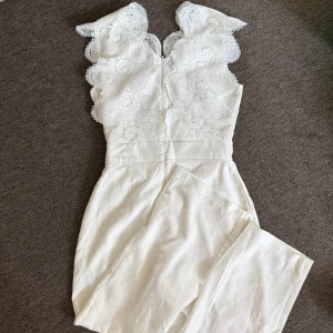 Vit spetsdetaljerad byxdress - Supersnygg vit byxdress med vida ben och hög midja. Överdelen har vackra spetsdetaljer och broderade mönster, vilket ger en romantisk och trendig look. Perfekt för dig som vill sticka ut med en elegant och unik outfit.