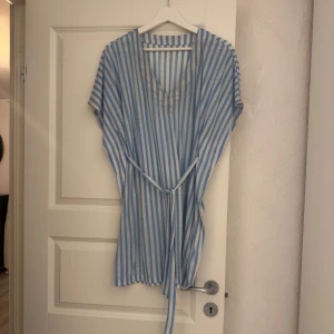 Blåvit randig strand klänning  - Superfin kort strand klänning i blåvitt randigt med v-ringning och knytband i midjan från H&M. Lätt och luftig modell med korta ärmar, perfekt över badkläder. Materialet känns som bomull eller linne. Då den är köpt för några år sedan säljer jag den billigt. 