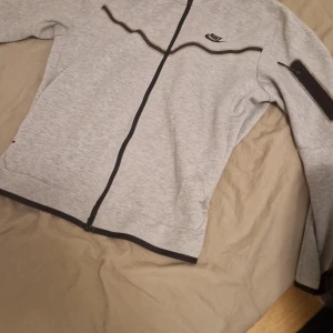 Grå Nike zipjacka med svarta detaljer - Snygg grå zipjacka från Nike med svarta detaljer och dragkedja framtill. Jackan har en svart Nike-logga på bröstet och en praktisk ficka med dragkedja på ärmen. Perfekt för en sportig och avslappnad stil.