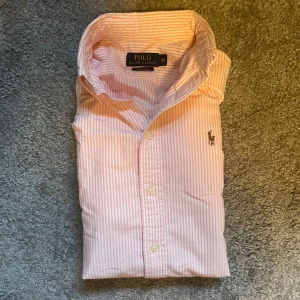 Rosa randig skjorta från Polo Ralph Lauren - Snygg rosa och vit randig skjorta från Polo Ralph Lauren i bomull. Klassisk krage, knappar framtill och broderad logga på bröstet. Custom fit-modell som sitter riktigt bra och passar perfekt till jeans eller chinos.