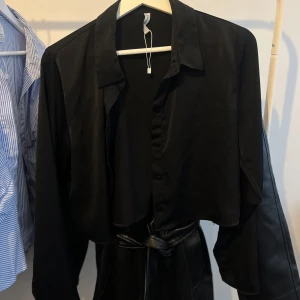 Svart croppad skjorta med knappar - Svart croppad skjorta med klassisk krage och långa ärmar. Skjortan har en snygg silkesliknande finish och knäppning framtill.