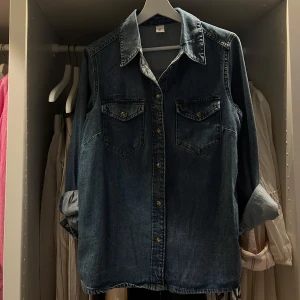 Blå jeansskjorta från H&M - Snygg blå jeansskjorta från H&M med klassisk krage, två bröstfickor med knappar och långa ärmar som kan rullas upp. Perfekt för dig som gillar en avslappnad och cool stil. Skjortan har tryckknappar framtill och är gjord i robust jeanstyg. Endast använd 1 gång på en bjudning.