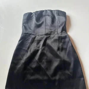 Svart axelbandslös klänning H&M - Snygg svart axelbandslös klänning från H&M med smockad rygg och slät framsida. Klänningen är kort och har en elegant, figurnära passform. Materialet är mjukt och har en lätt glansig yta som ger en lyxig känsla. Perfekt för dig som vill ha en stilren look.