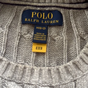 Grå stickad tröja från Polo Ralph Lauren - Notera att det är en M  (10-12) passar min son som  14 och 165.  grå kabelstickad tröja från Polo Ralph Lauren med rund hals och ribbade muddar. Tröjan har lång ärm och den klassiska Polo-loggan broderad på bröstet. Perfekt för dig som gillar stilrena och tidlösa plagg.