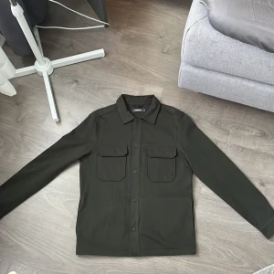 Mörkgrön overshirt  - Snygg mörkgrön overshirt med klassisk krage och två stora bröstfickor med lock. Rak passform och långa ärmar, tillverkad i ett slitstarkt bomullsmaterial. Stängs med knappar framtill och har en stilren, minimalistisk look. Använd ett fåtal gånger