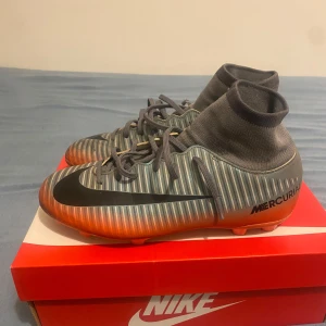 Nike Mercurial randiga fotbollsskor - Snygga Nike Mercurial fotbollsskor med grå och silverrandigt mönster, svarta detaljer och orange sula. Skorna har hög strumpliknande krage och snörning för extra support. Perfekta för dig som vill sticka ut på planen.