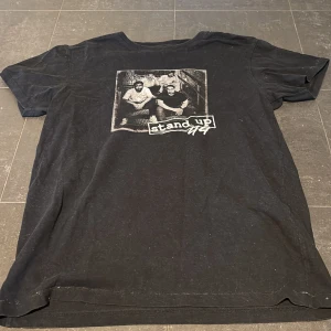 T-shirt - Svart t-shirt med stort grafiskt tryck på bröstet som visar fyra personer sittande på en trappa och texten 'stand up'. T-shirten har rund hals och korta ärmar. Materialet känns som mjuk bomull och passformen är klassisk och avslappnad.