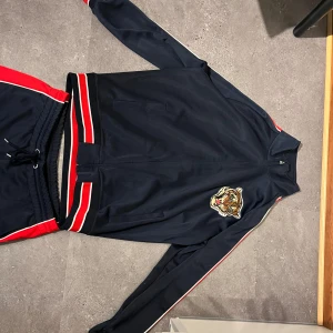 GANT blå och röda mjukis set L - Snygga mjukisbyxor från GANT i marinblått med röda sidopaneler och vita detaljer. Byxorna har elastisk midja med snörning och relaxed fit. Perfekta för chill dagar eller träning, matcha gärna med tillhörande jacka för en komplett look.