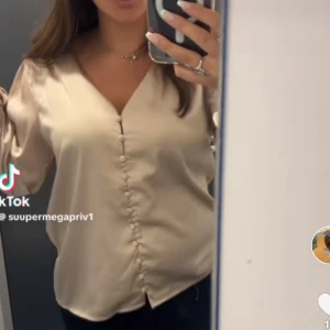 Champagnefärgad blus med knappar - Säljer en champagnefärgad blus med v-ringning och dekorativa knappar framtill. Blusen har långa ärmar och ett mjukt, glansigt material som ger en lyxig känsla. Perfekt för dig som gillar stilrena och eleganta plagg.