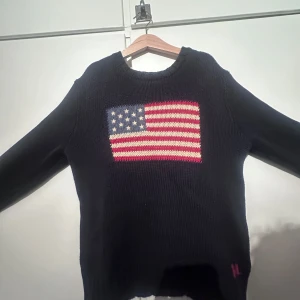 Mörkblå stickad tröja Polo Ralph Lauren - Storlek: M(barn) 10-12 år. Snygg mörkblå stickad tröja från Polo Ralph Lauren med amerikansk flagga på bröstet. Klassisk rund halsringning och långa ärmar. Perfekt för dig som gillar preppy stil och vill sticka ut med en ikonisk detalj framtill.