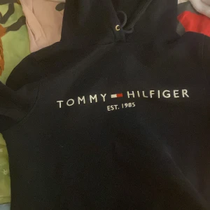 Svart hoodie Tommy Hilfiger S - Svart hoodie från Tommy Hilfiger i storlek S med klassisk logga och texten 'EST. 1985' broderad på bröstet. Tröjan har huva med snörning och är perfekt för en chill och stilren look. Säljer för att jag aldrig använt den❤️