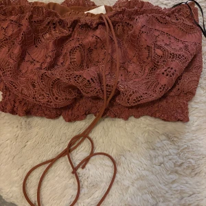 Rostbrun spets bandeau tubtopp H&M - Superfin bandeau tubtopp från H&M i rostbrun färg med detaljerad spets och snörning framtill. Toppen är axelbandslös och har en croppad passform som ger en snygg siluett. Perfekt för dig som vill sticka ut med coola detaljer och unik look.