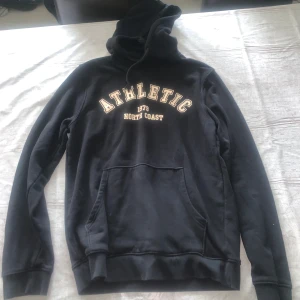 Svart hoodie Athletic North Coast - Svart hoodie med stor, mjuk huva och snörning. Framtill står det 'ATHLETIC 1979 NORTH COAST' i beige bokstäver. Tröjan har en klassisk känguruficka och långa ärmar. Perfekt för chill dagar eller när du vill ha en sportig look.