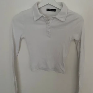 Vit ribbad pikétröja från New Yorker  - Vit pikétröja från New Yorker i ribbad bomull med klassisk krage och tre knappar framtill. Långärmad och croppad passform som ger en fräsch och clean look. Perfekt för dig som gillar stilrena och enkla plagg med lite edge.