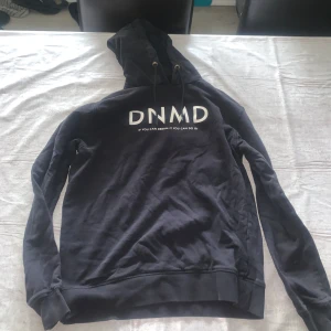 Svart hoodie från DNMD med tryck - Svart hoodie från DNMD med vit texttryck på bröstet och snörning i huvan. Tröjan har långa ärmar och en klassisk passform, perfekt för chill dagar. Materialet känns mjukt och bekvämt mot huden. Storlek L, men passar M