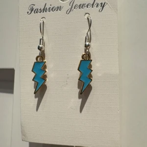 Blå blixtformade örhängen - Coola örhängen i form av blixtar med blå färg och guldfärgade kanter. Perfekt för dig som vill sticka ut med en unik och edgy stil. Hängande modell med metallkrok för enkel användning.