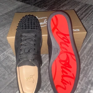 Christian Louboutin  - Svarta sneakers från Christian Louboutin i mocka med ikonisk röd sula och svarta nitar på tån. Skorna har snörning och rund tå, samt exklusiva detaljer som gör dem till ett statement. Perfekta för dig som vill sticka ut med lyxig streetstyle.