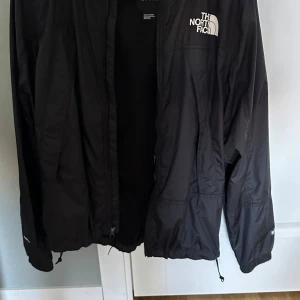 Svart vind/regnjacka från The North Face - Svart vindjacka från The North Face med dragkedja framtill, justerbar nederkant med snörning och meshfoder. Jackan har lång ärm, diskret logga på bröstet och är tillverkad i lätt polyester. Perfekt för blåsiga dagar .