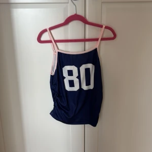Mörkblå linne från SHEIN med siffra 80 på  - Sportig mörkblå linne från SHEIN med vita siffran 80 framtill och ljusrosa tunna axelband. Toppen har en enkel, rak passform och är perfekt för en avslappnad look. Materialet är mjukt och stretchigt, vilket gör den bekväm att bära hela dagen. Den är även aldrig använd. 