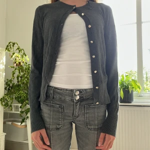 Mörkgrå kofta med silvriga knappar - Snygg mörkgrå kofta med rund hals och guldiga knappar framtill. Koftan har långa ärmar med breda ribbade muddar och är gjord i ett mjukt stickat material. Perfekt att slänga över en t-shirt för en chill look. Passar dig som gillar enkel och stilren design.