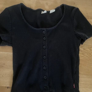 Svart ribbad topp från Levi's XS - Svart ribbad topp från Levi's med kort ärm och knappar hela vägen framtill. Djup rundad urringning och figurnära passform. Klassisk röd Levi's-tag i sidan. Perfekt att styla med jeans eller kjol för en cool vardagslook.