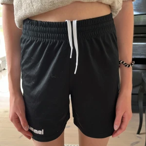 Svarta träningsshorts från Hummel - Svarta Hummel shorts med vit snörning i midjan och vita chevron-detaljer längs sidan. De har elastisk midja och är gjorda i ett lätt, syntetiskt material som passar perfekt för sport och träning. Snygg och sportig look med normal passform.