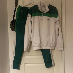 Lacoste tracksuit  - Säljer ett par snygga träningsbyxor från Lacoste i grönt och vitt. Byxorna har resår i midjan, raka ben och klassisk Lacoste-logga framtill. Materialet är lätt och syntetiskt, perfekt för sport eller chill. Matchar jackan i samma set för en clean retrostil.