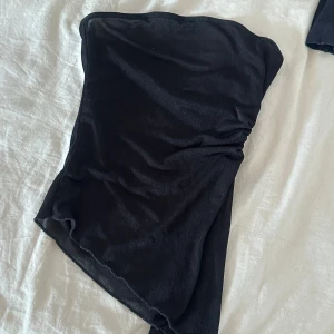 Svart bandeau tubtopp med knyt - Säljer en svart bandeau tubtopp med snörning och knyt i sidan. Toppen har en asymmetrisk form och är gjord i ett tunt, stretchigt material som känns mjukt mot huden. Perfekt för dig som vill ha en stilren och trendig look.